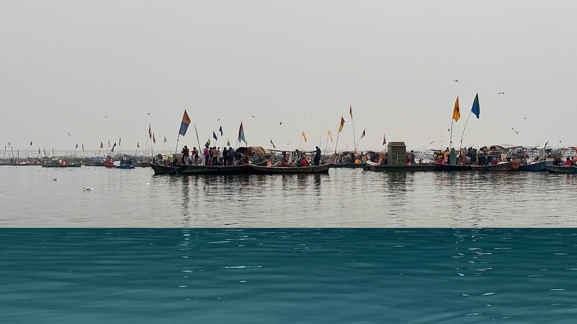 Scenic photograph capturing the confluence of two rivers, Yamuna and Ganga, featuring numerous shallow boats along the horizon marked by red, yellow, and blue flags, with people standing and sitting on them engaged in ritual activities